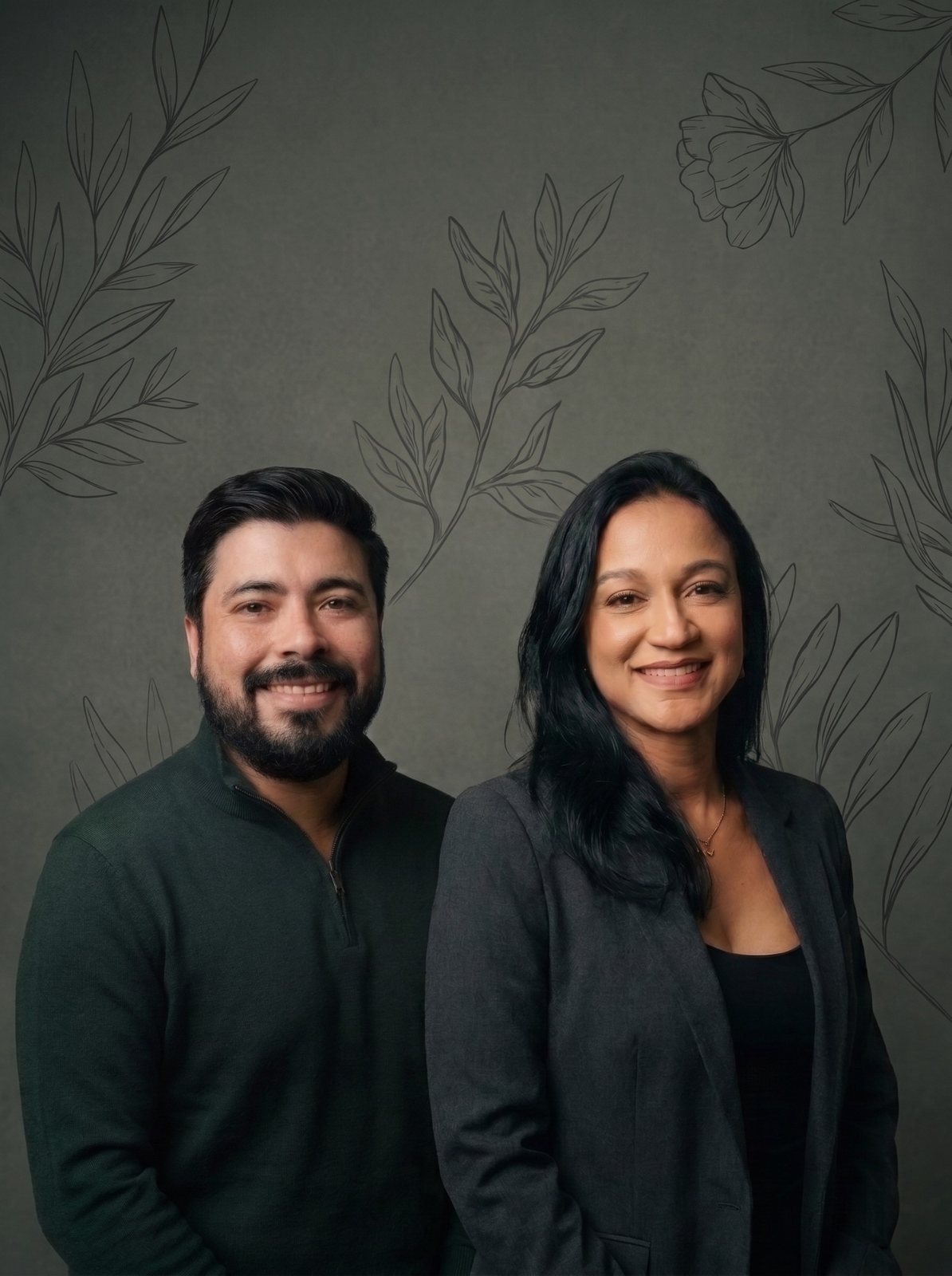 Denis Faria e Ana Oliveira, lado a lado e sorrindo, diante de uma parede verde-escura com ilustrações de folhas.