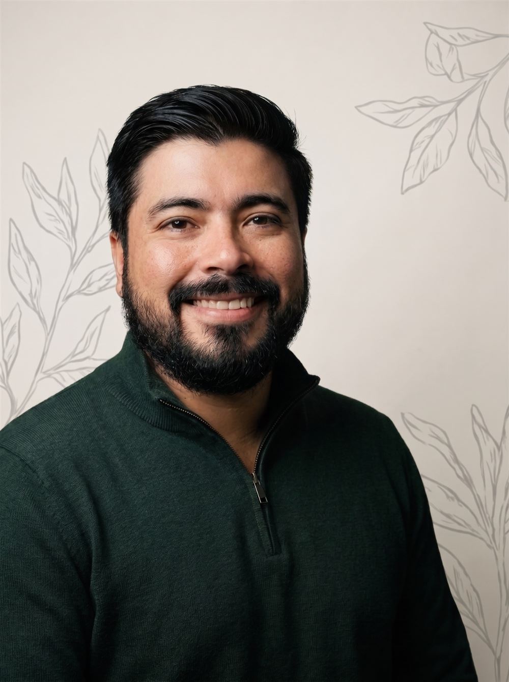 Retrato de Denis Faria, sorrindo, de suéter verde-escuro, diante de parede creme com desenhos de folhas